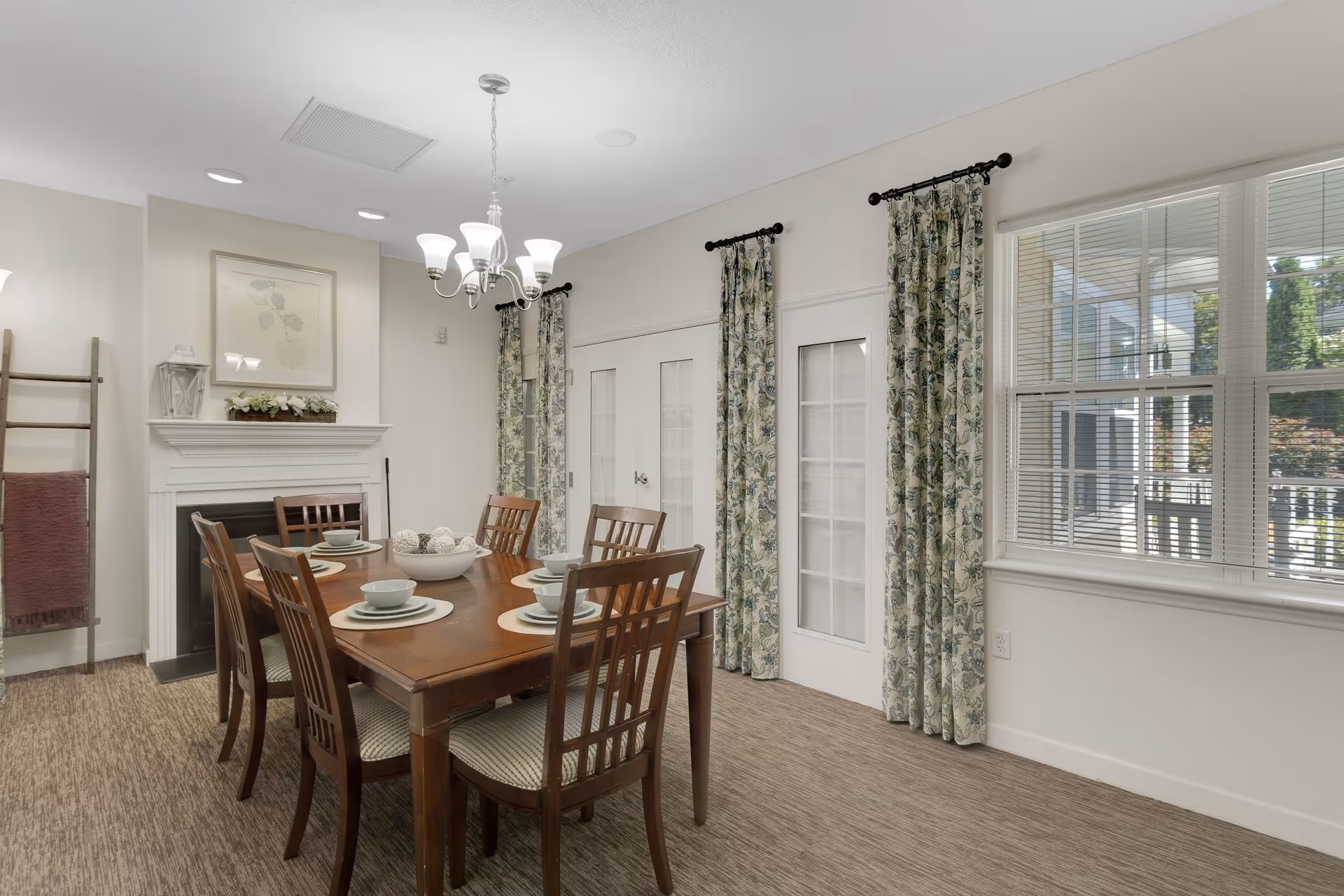A dining room with a wooden table set for six with plates, bowls, and cups. The room features a white fireplace with decorative items on the mantel, floral patterned curtains on the windows and glass-paneled doors, and a chandelier hanging above the table. The walls are painted light beige, and the floor is carpeted.