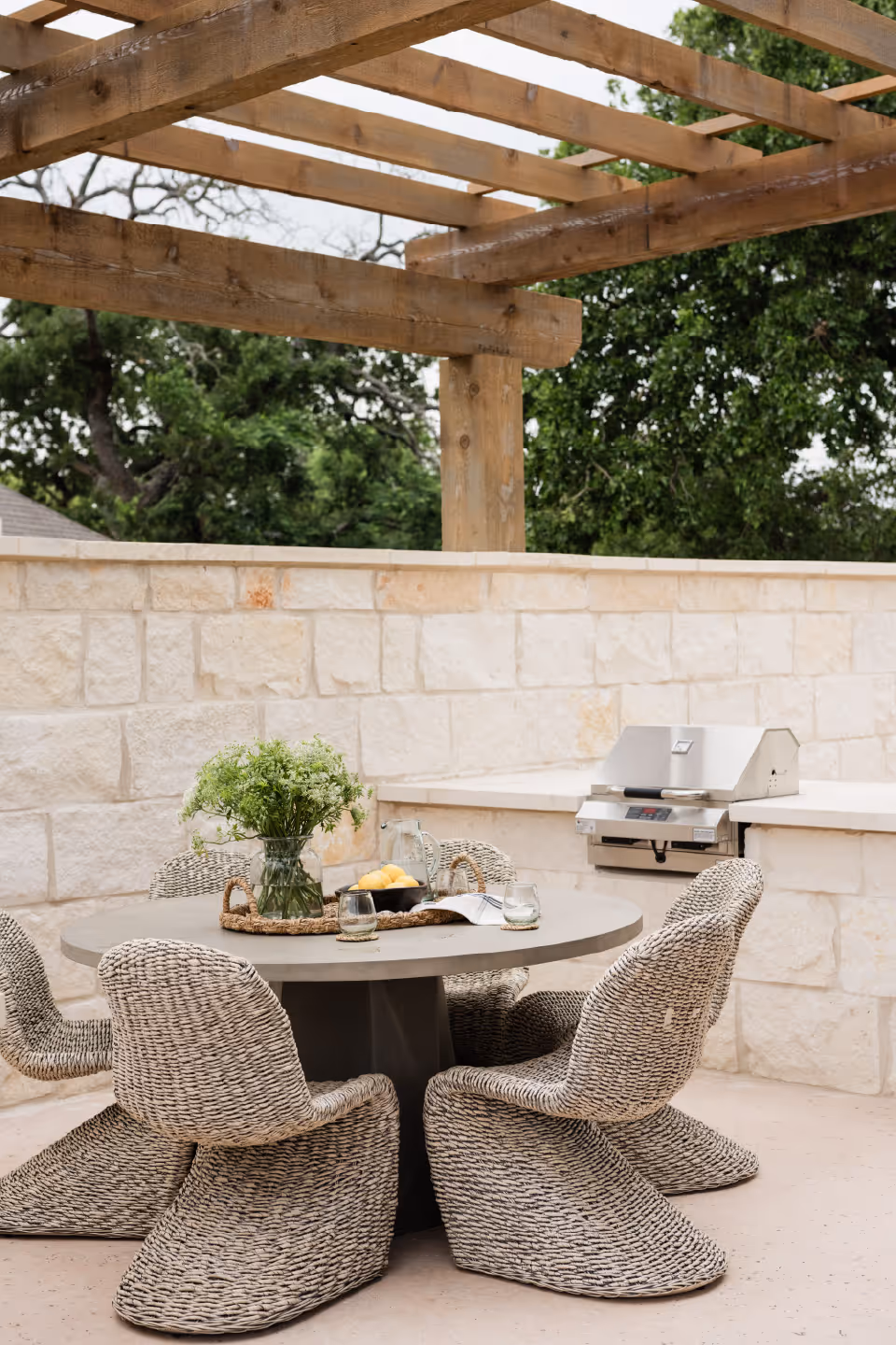 Outdoor patio area with a round table surrounded by four woven chairs. On the table, there is a tray with a vase of green flowers, a bowl of lemons, and two glasses. In the background, there is a built-in stainless steel grill and a stone wall with wooden pergola beams overhead. Trees are visible beyond the wall.