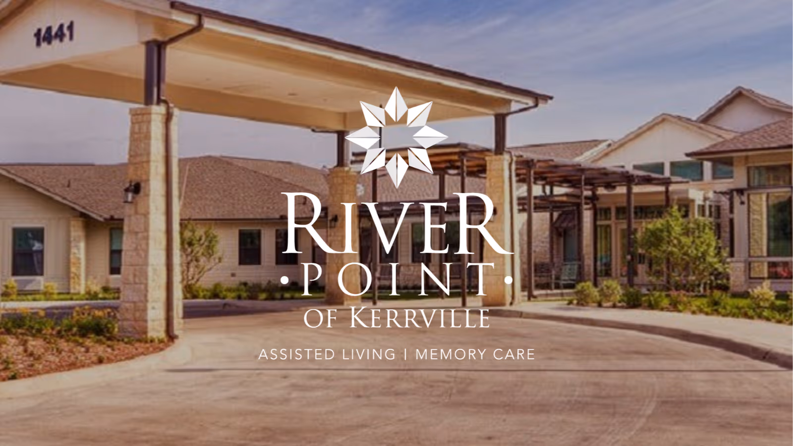 Exterior view of River Point of Kerrville assisted living and memory care facility showing a covered entrance with stone pillars, a driveway, and surrounding buildings with landscaped greenery.