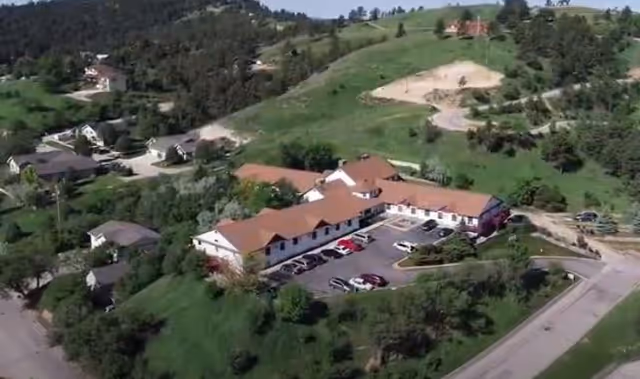 Aerial view of a single-story building with a red roof surrounded by greenery and hills, with a parking lot containing several cars in front of the building.