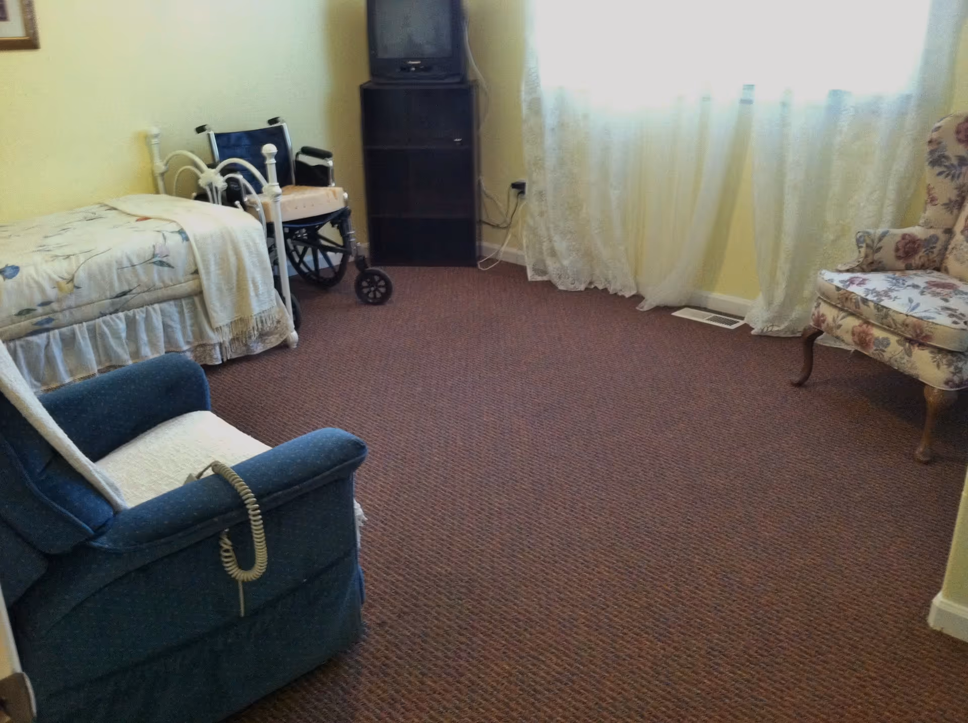 A cozy bedroom with a single bed covered in a floral bedspread, a blue recliner chair with a white cushion and a corded remote, a wheelchair beside the bed, a small dark wooden TV stand with an old-style TV on top, and a floral upholstered armchair near a window with sheer white curtains.
