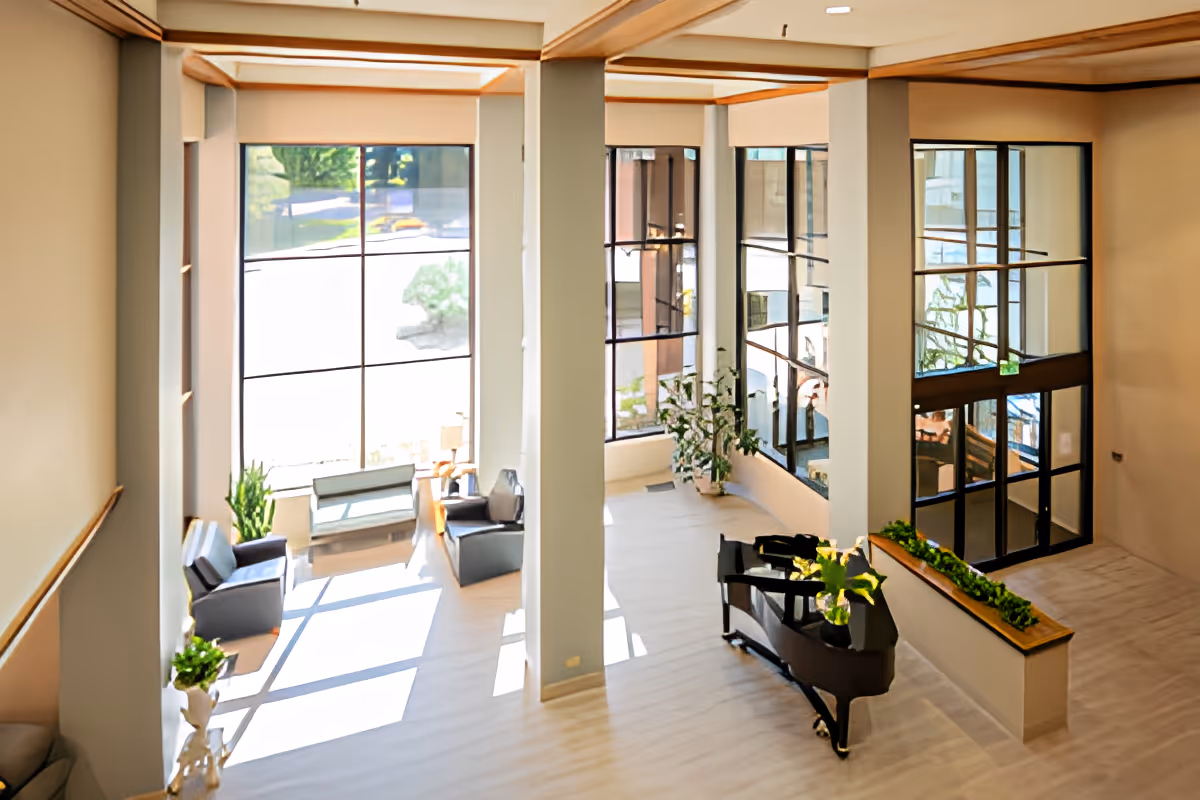 Bright and spacious interior common area with large floor-to-ceiling windows letting in natural light. The room features several seating arrangements with chairs and benches, a grand piano with plants on top, and decorative plants near the windows and columns.