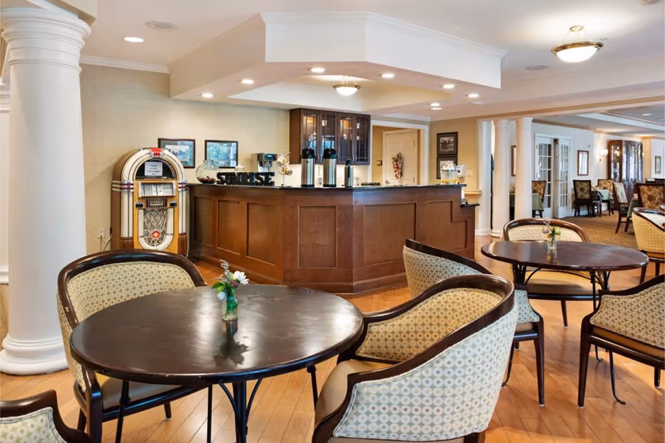 A cozy common area in a senior living facility featuring round wooden tables with patterned cushioned chairs, a wooden counter with coffee dispensers, a vintage jukebox, and decorative columns. The space is warmly lit with ceiling lights and has framed pictures on the walls.