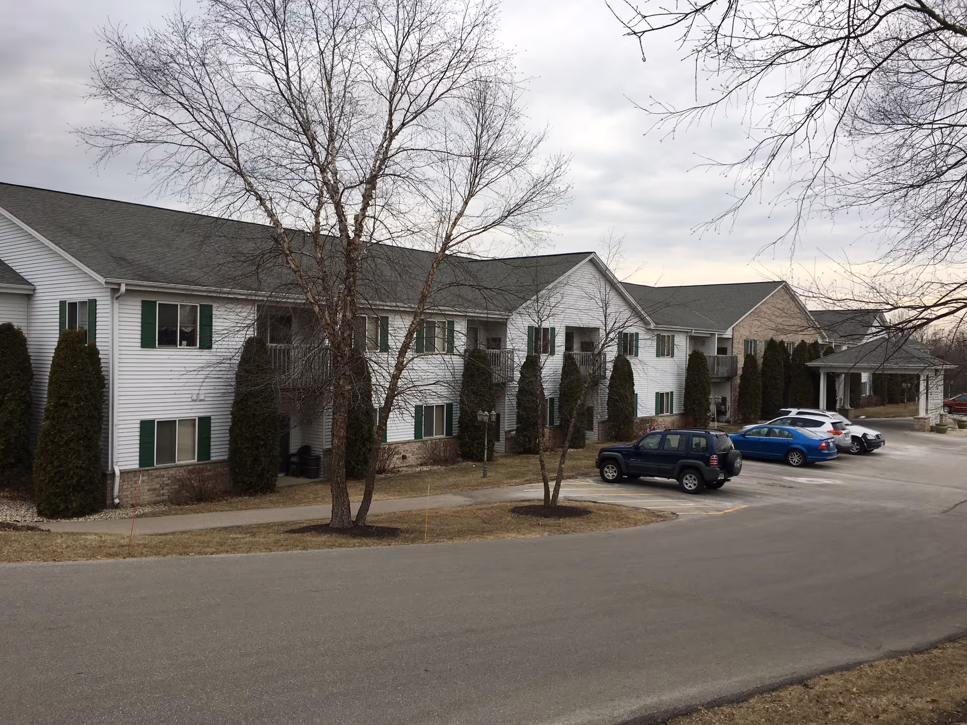Exterior view of a two-story senior living apartment building with white siding and green shutters. Several tall evergreen shrubs and leafless trees are in front of the building. There are a few parked cars along the driveway and a covered entrance on the right side.