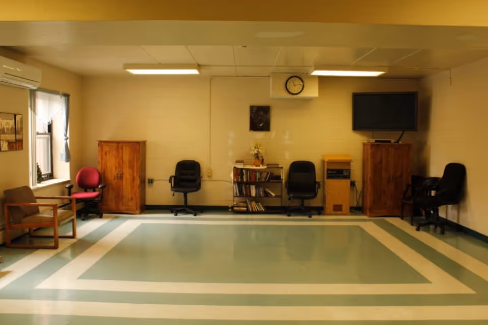 A spacious activity room with chairs, wooden cabinets, a bookshelf, a wall clock and a TV over a patterned linoleum floor.