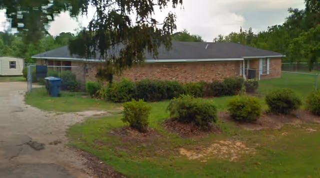 Single-story brick building with a dark shingled roof surrounded by green bushes and grass. There is a dirt driveway on the left side with two blue trash bins near the building. Trees and a cloudy sky are visible in the background.