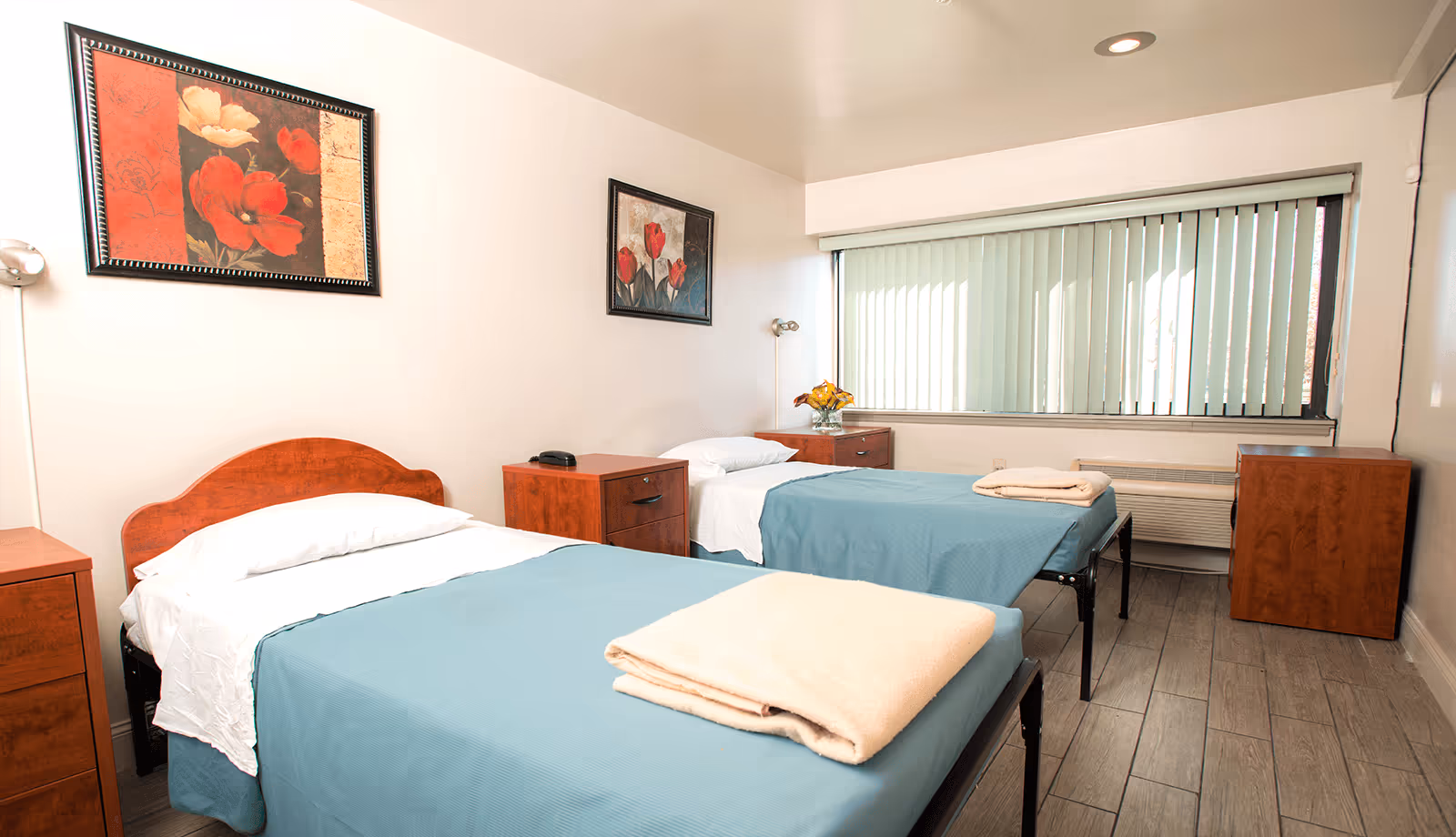 A simple, clean bedroom with two single beds covered in blue bedspreads and white sheets. Each bed has a folded beige blanket at the foot. There are wooden nightstands between and beside the beds, one with a telephone and the other with a vase of yellow flowers. The room has light-colored walls adorned with two framed floral paintings and a large window with vertical blinds letting in natural light. The floor is covered with wood-like tiles.