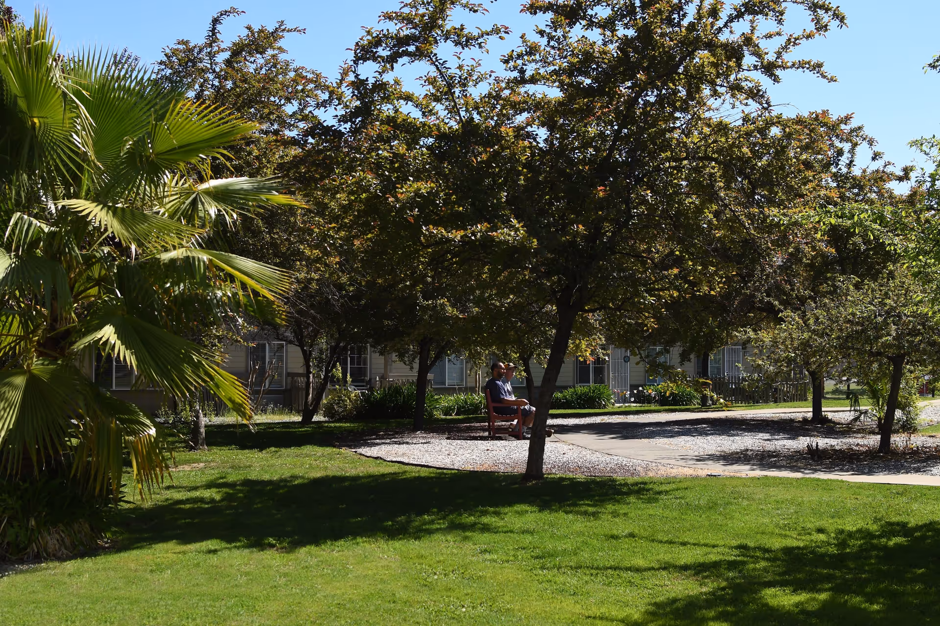 A peaceful garden area at Oakdale Heights Assisted Living, featuring a green lawn, trees, and a bench where two individuals are seated, enjoying the outdoors.