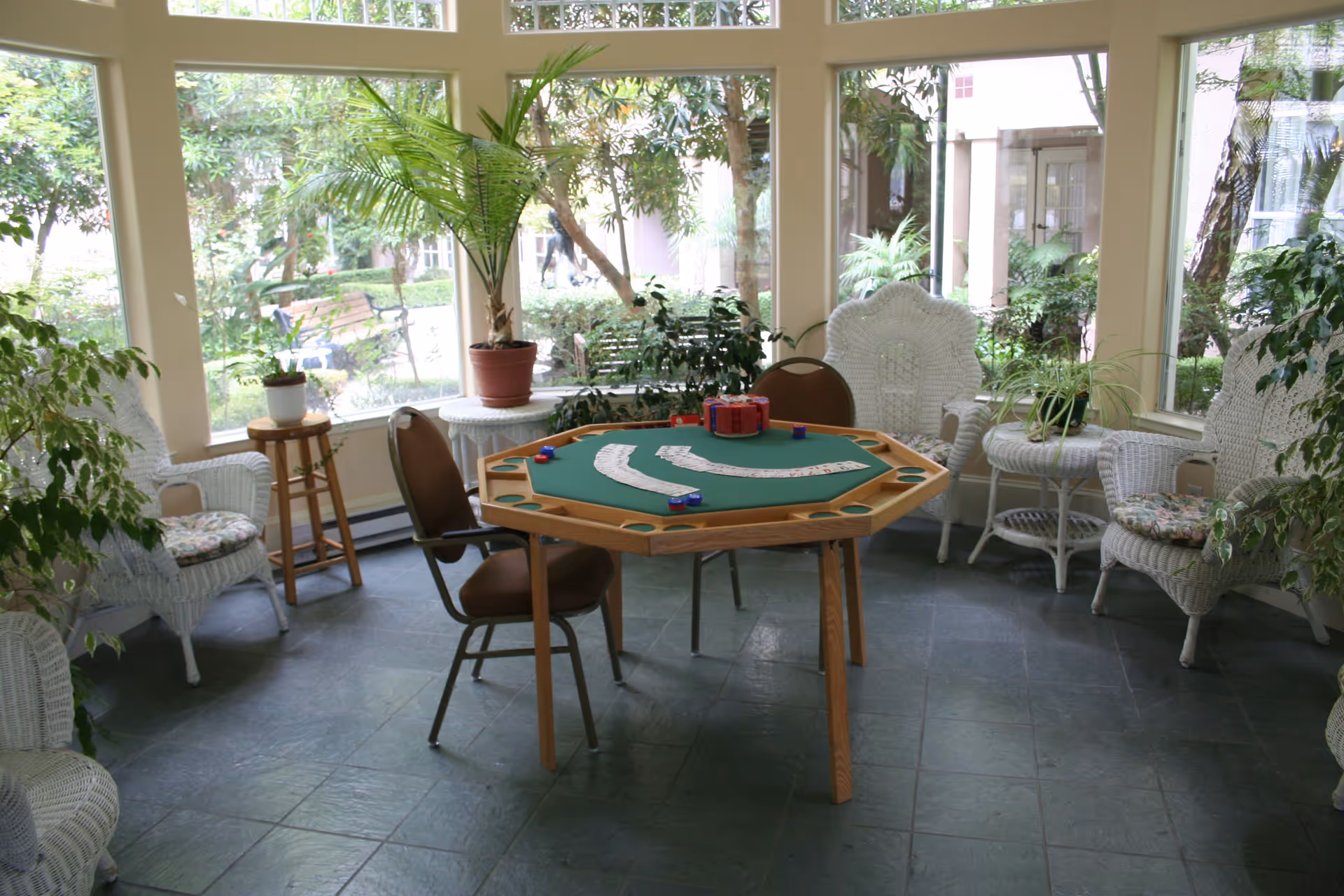 A sunlit room with large windows showing a garden outside. Inside, there is a green felt octagonal game table with playing cards and poker chips on it. Surrounding the table are several chairs, including white wicker chairs with floral cushions and brown chairs. Various potted plants are placed around the room, creating a cozy and inviting atmosphere.