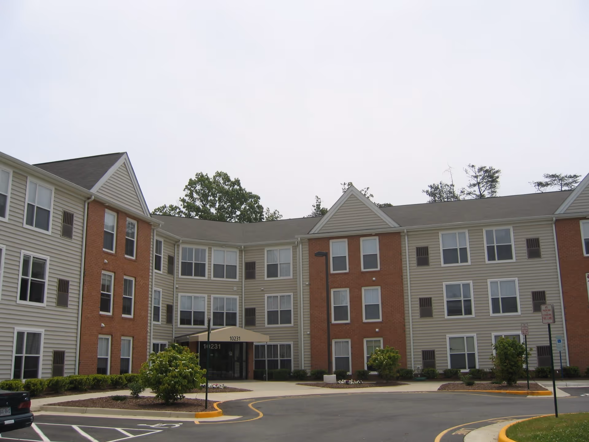 Exterior view of a three-story senior living facility building with beige siding and red brick accents, multiple windows, and a covered entrance with the number 10231 above the door. There are small landscaped areas with bushes and a paved driveway in front.