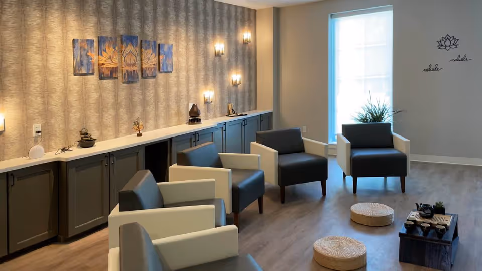 A modern senior living common area with six cushioned armchairs arranged in a semi-circle around two round woven floor cushions and a low wooden table with tea set. The room features a long cabinet with decorative items and wall-mounted lights, a large window with a plant in front, and wall art including a lotus flower painting and the words 'exhale inhale' with a lotus flower decal.