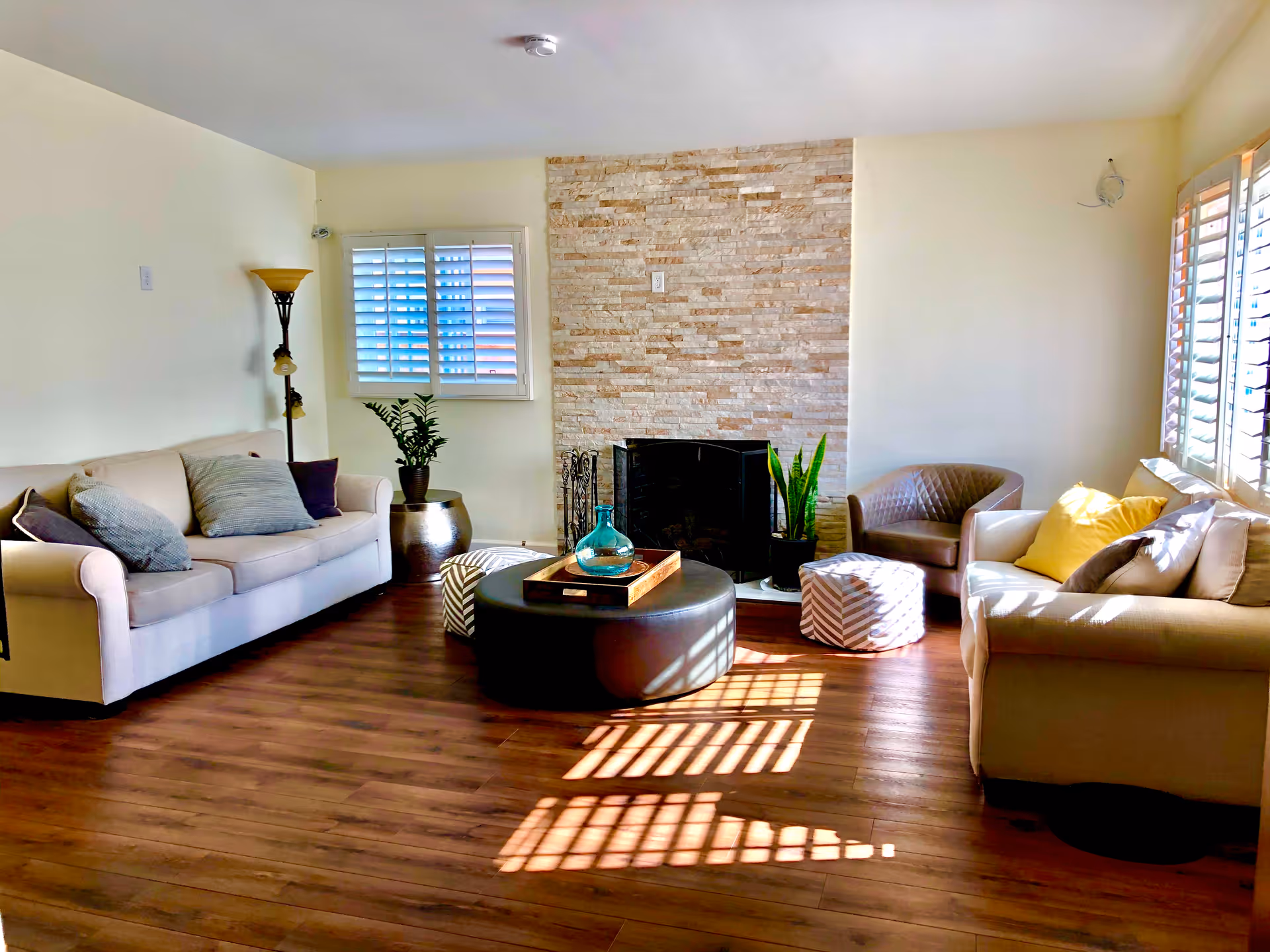 A bright living room with two beige sofas, a round black ottoman with a wooden tray and glass vase on top, two patterned poufs, a brown armchair, a tall floor lamp, and a stone fireplace. The room has wooden flooring and windows with white shutters letting in sunlight.
