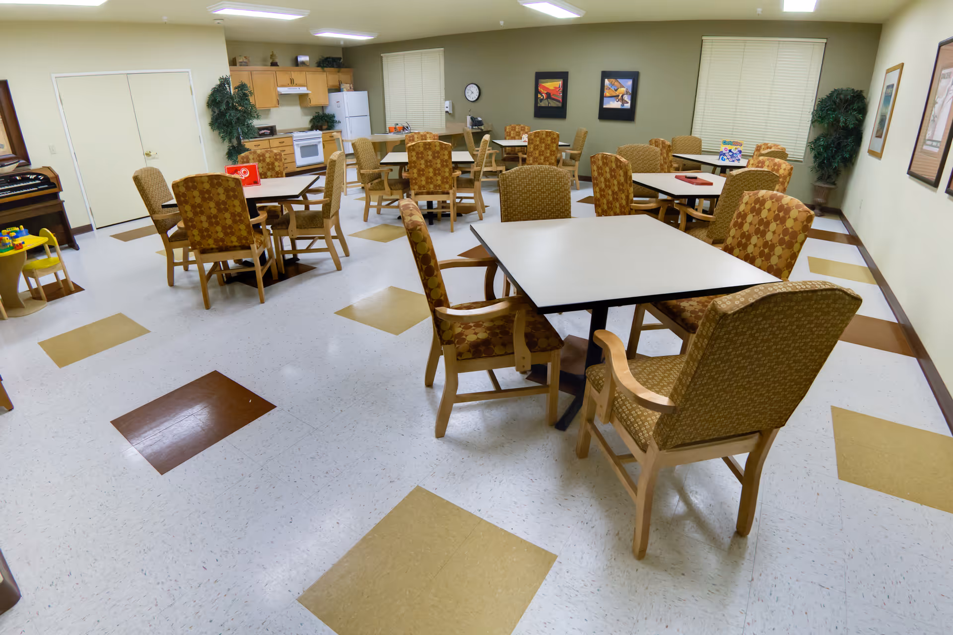 Communal dining/activity room with several square tables, patterned armchairs, a kitchenette and a piano in the background.