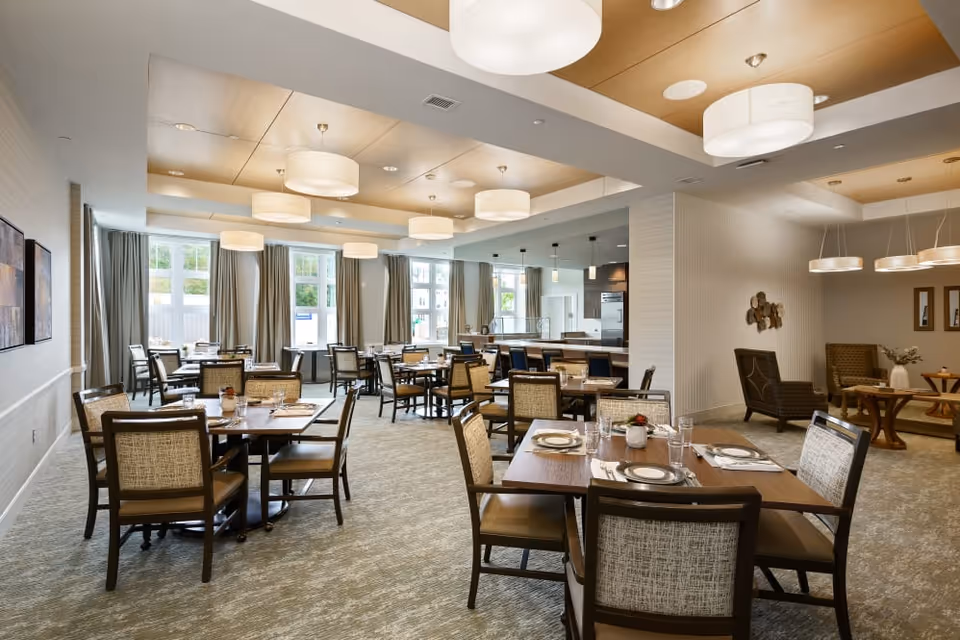 A spacious and well-lit dining room with multiple wooden tables and cushioned chairs arranged neatly. Each table is set with plates, glasses, and napkins. Large windows with curtains allow natural light to fill the room. Modern ceiling lights hang from a wooden paneled ceiling. In the background, there is a kitchen area with bar stools and a cozy seating area with armchairs and small tables.