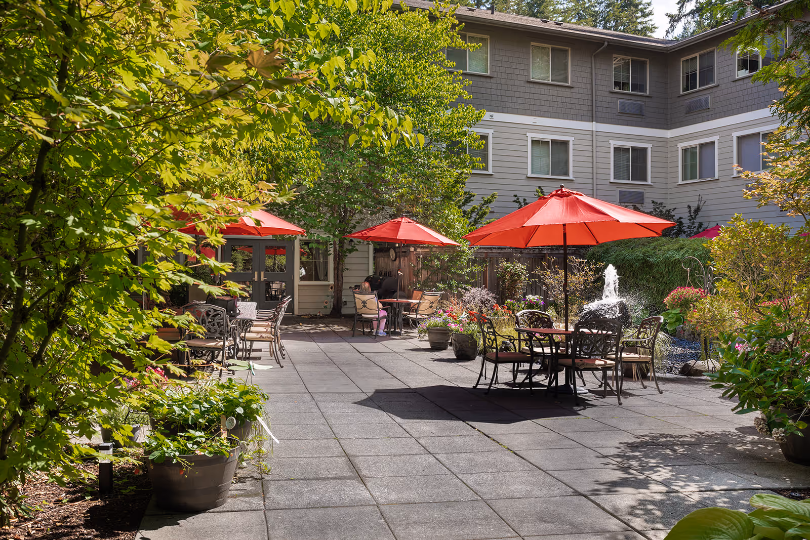 Outdoor courtyard area at Village Concepts of Issaquah-Spiritwood at Pine Lake with several tables and chairs under red umbrellas, surrounded by greenery and potted plants. A water fountain is visible near the center right, and the building with multiple windows forms the background.