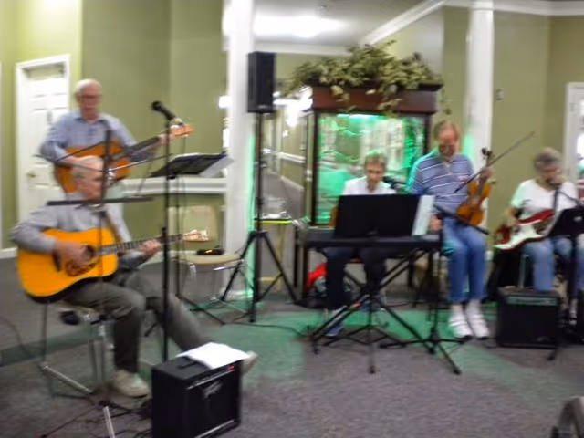 A group of five elderly individuals playing musical instruments indoors. Two are playing acoustic guitars, one is playing a keyboard, one is playing a violin, and one is playing an electric bass guitar. They are seated in a room with green walls and a large fish tank in the background.