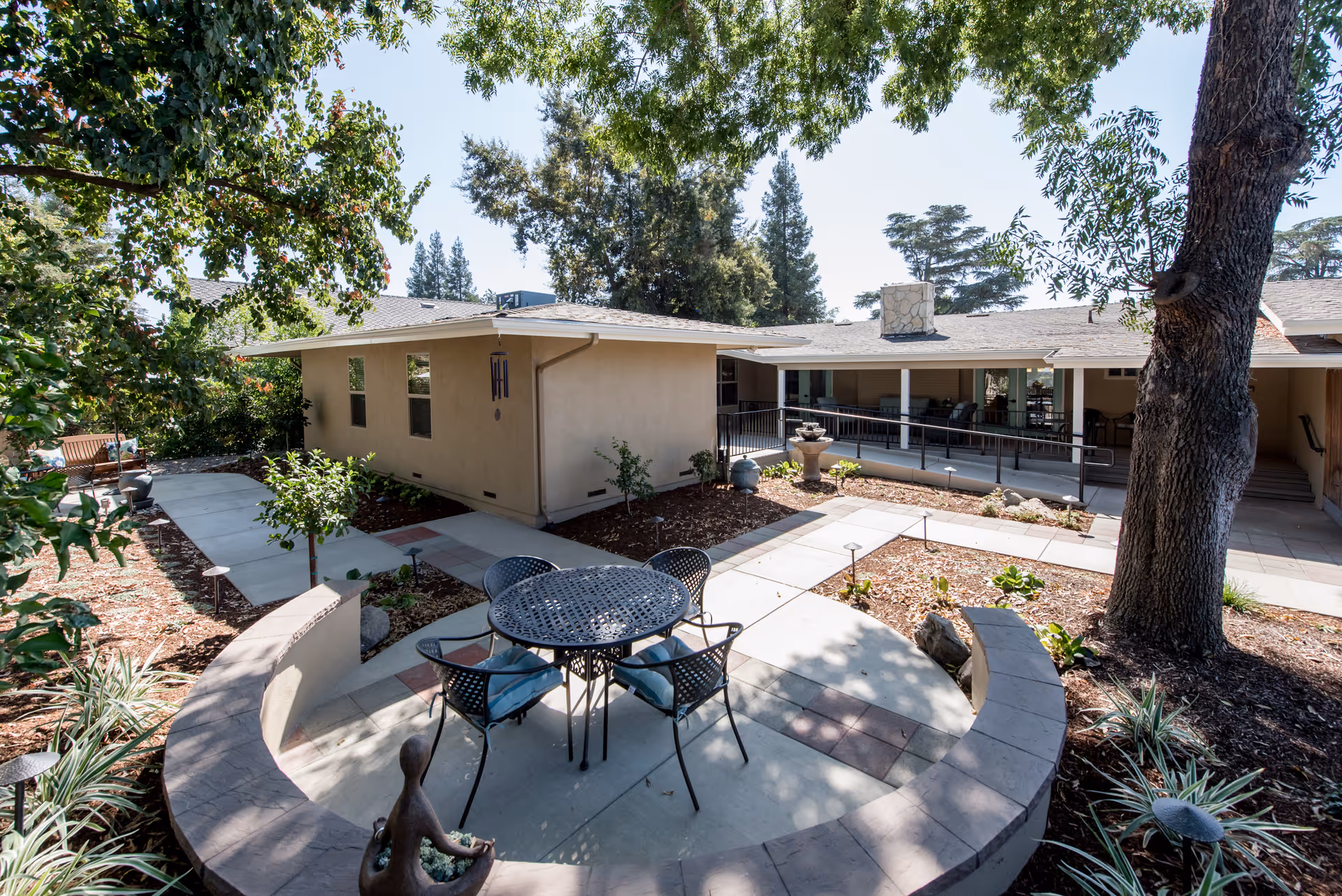 Outdoor courtyard area at Kaweah Health Ruth Wood Open Arms House featuring a round metal table with four chairs with cushions, surrounded by a curved stone bench. The courtyard is landscaped with plants, trees, and a small fountain near the building. The building has a covered patio with seating and a stone chimney, and there are paved walkways throughout the garden area.