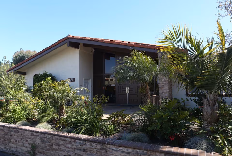 Entrance of a single-story stucco building with a red-tiled roof, double doors and lush palm landscaping behind a low stone wall.
