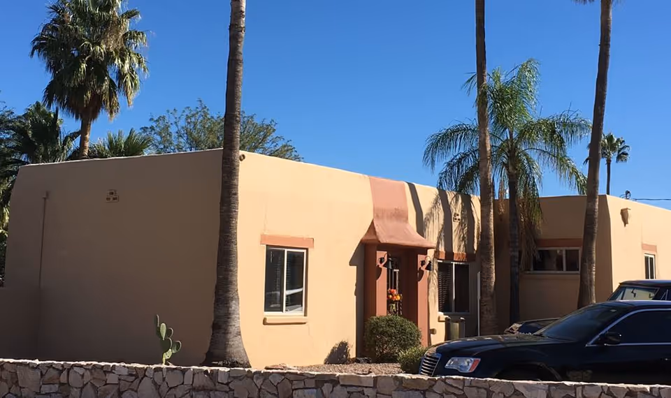 Single-story stucco Southwestern-style building front with palm trees, a low stone wall and parked cars under a clear blue sky.