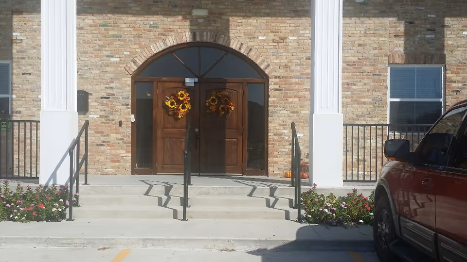 Front entrance of a brick building with double wooden doors adorned with wreaths, white columns, and steps leading up from a parking area.