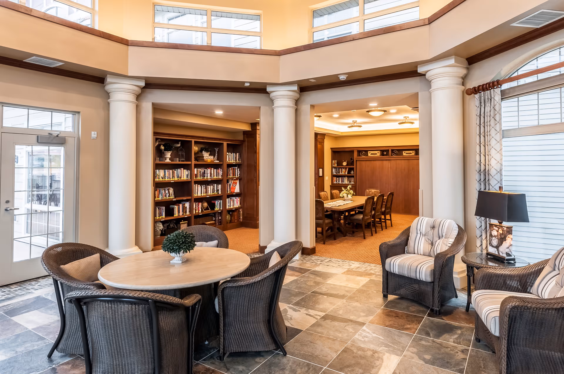 A bright and spacious common area in a senior living facility featuring a round table with four wicker chairs and a small plant centerpiece. The room has large windows with white blinds and beige walls with white columns. In the background, there is a cozy library area with bookshelves filled with books and a long wooden table surrounded by chairs. The floor is tiled with large stone tiles.