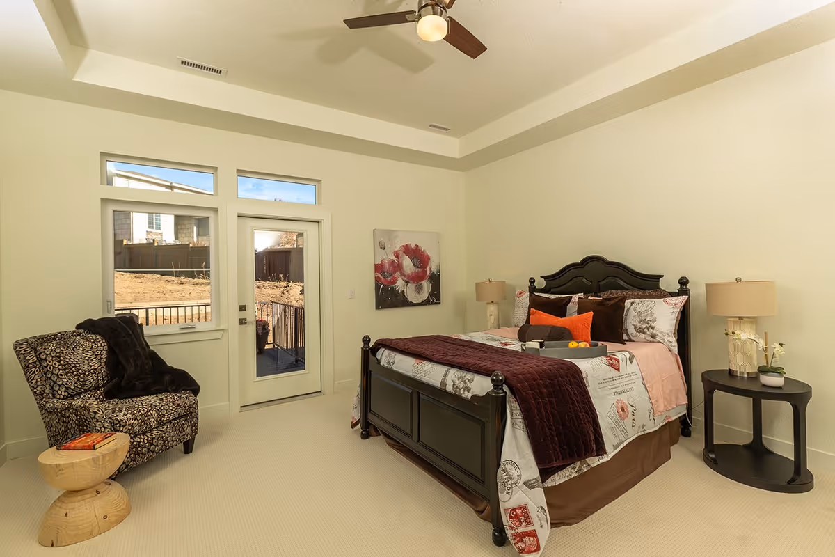 A cozy bedroom with a dark wooden bed frame, floral and patterned bedding, and multiple pillows. There is a small round side table with a lamp and a plant on the right side of the bed. On the left side of the room, there is a patterned armchair with a dark throw blanket and a small wooden stool with a book on it. The room has a ceiling fan and a door with windows above it leading outside, allowing natural light to enter.