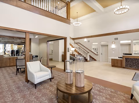 Spacious senior living facility lobby with seating, a reception desk, staircase, and high coffered ceilings.