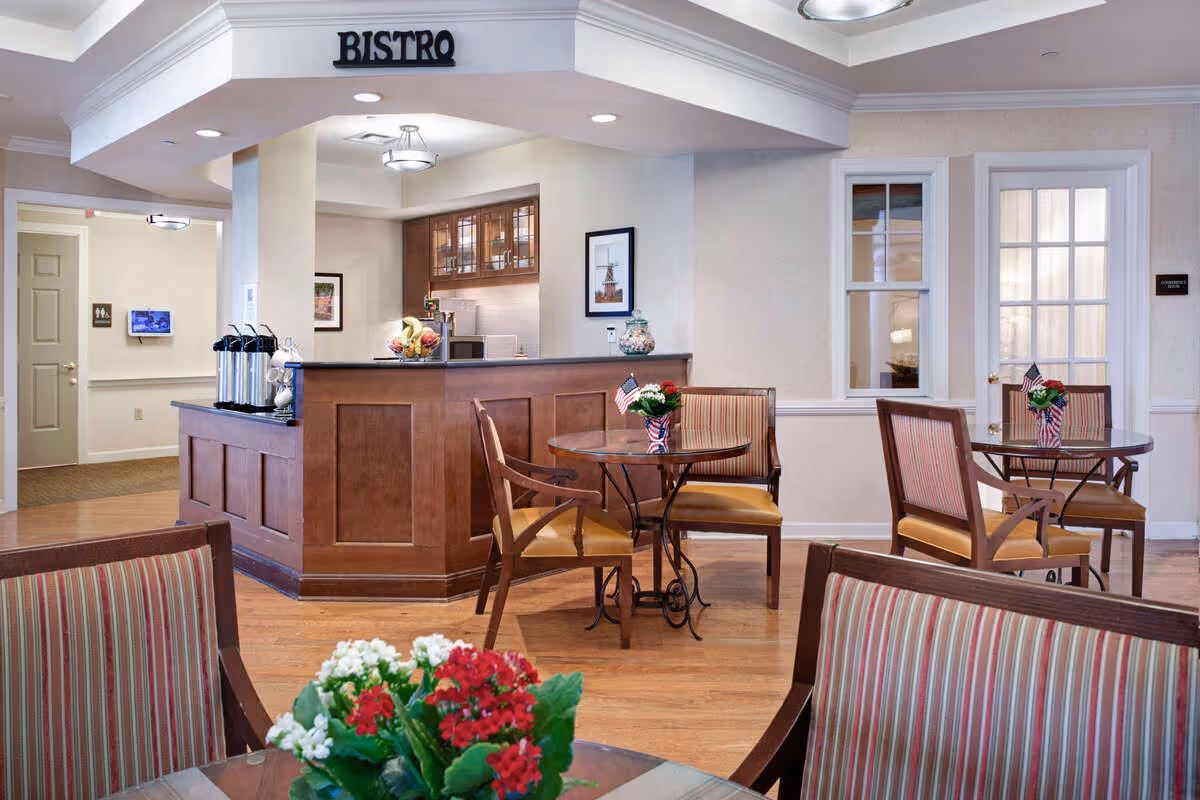 Interior view of a bistro area in a senior living facility with wooden tables and chairs, small American flag decorations, and flower arrangements on the tables. The bistro counter is visible with coffee dispensers and fruit on the counter. The room has light-colored walls, wooden flooring, and ceiling lights.