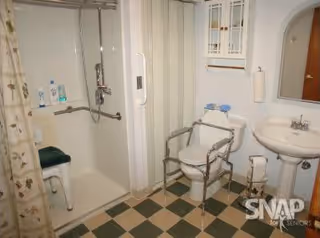 Bathroom with a shower area featuring a grab bar and shower seat, a toilet with safety rails, a pedestal sink with a mirror above it, and a checkered floor pattern.