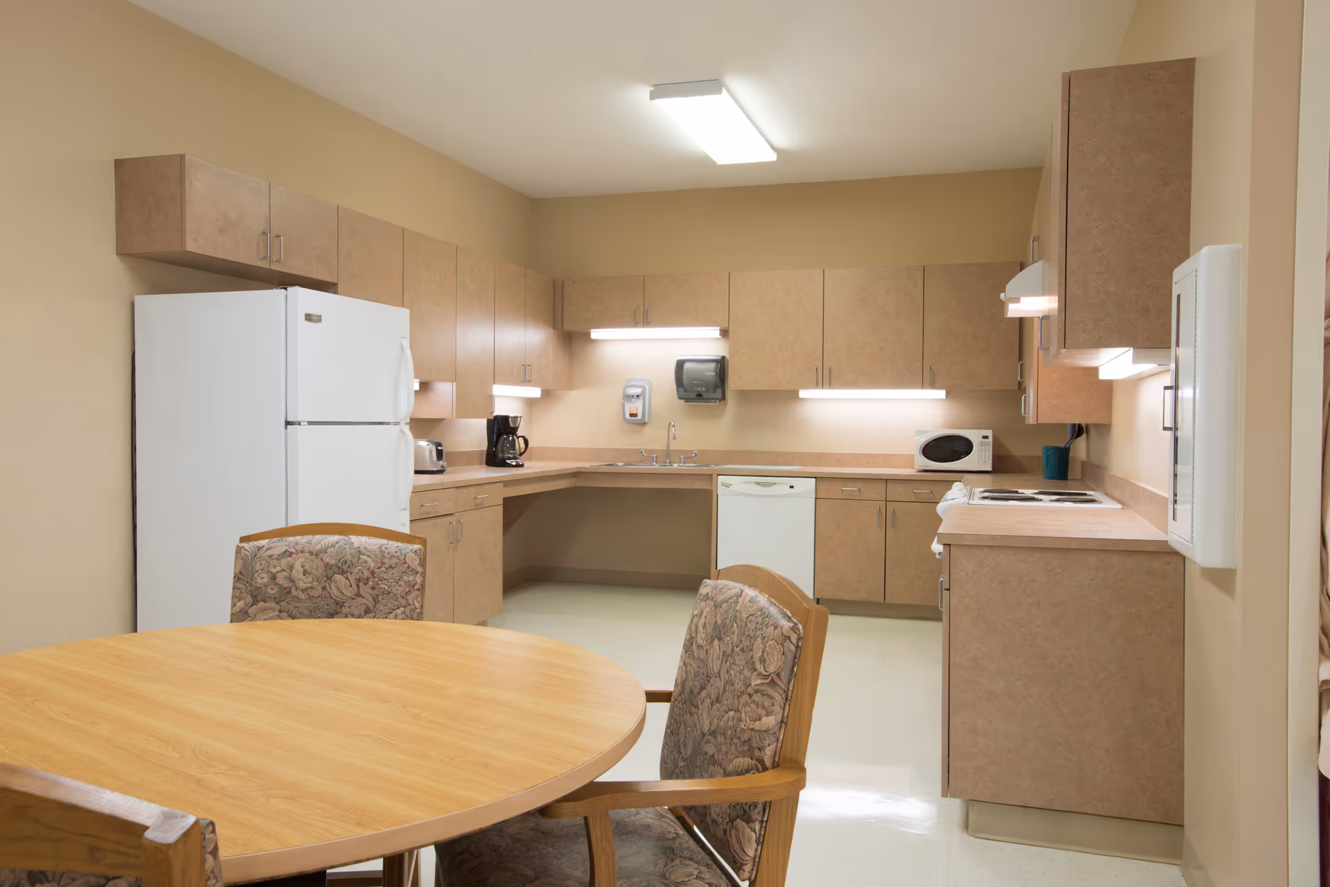A clean and well-lit kitchen area with beige cabinets, a white refrigerator, dishwasher, microwave, coffee maker, toaster, and a round wooden table with floral upholstered chairs.