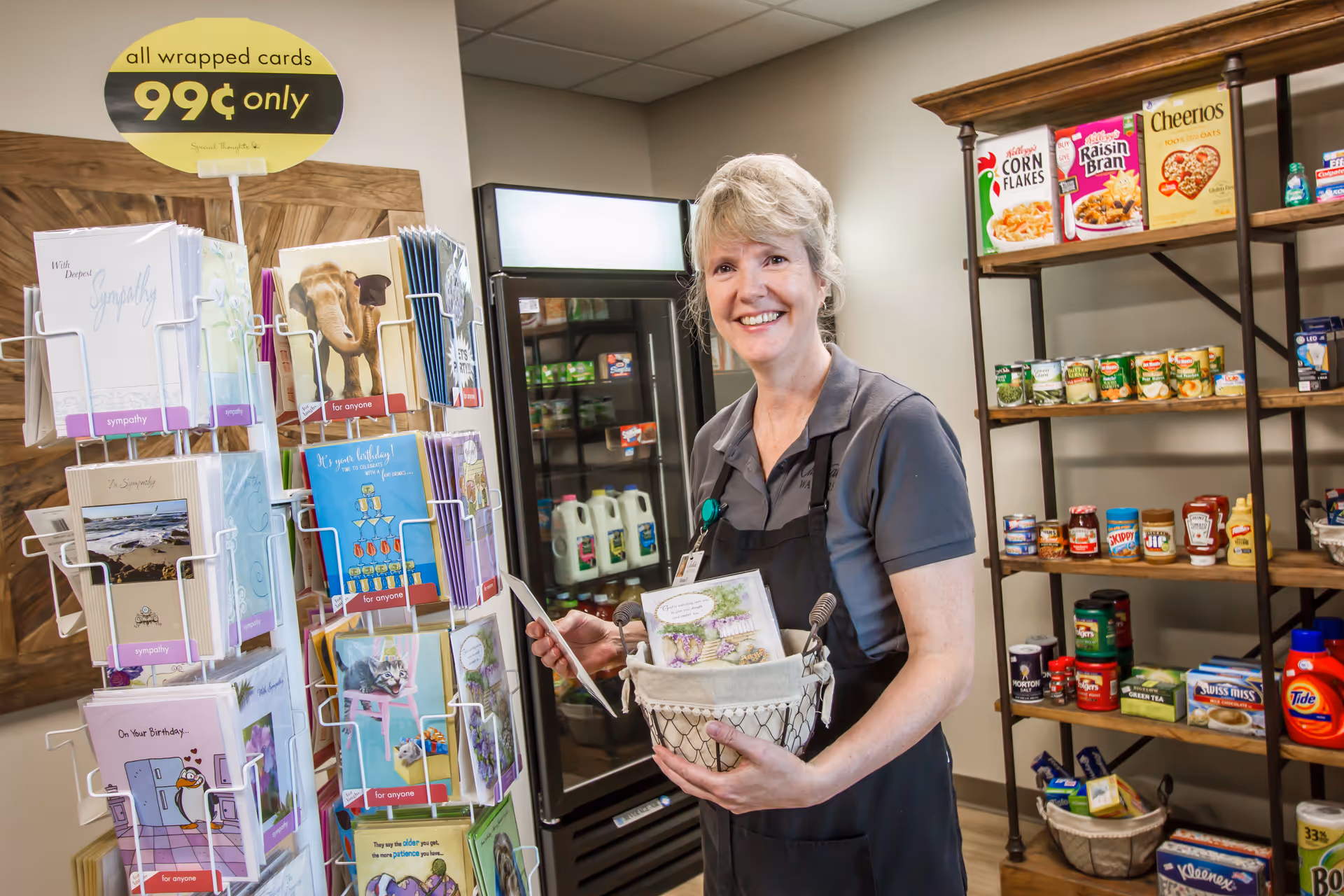 A smiling woman wearing a dark apron stands in a small shop area holding a basket filled with greeting cards. To her left is a rotating rack filled with various greeting cards priced at 99 cents each. Behind her are shelves stocked with groceries including cereal boxes, canned goods, and household items like laundry detergent. A refrigerator with milk and other items is also visible in the background.