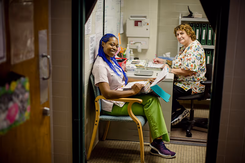 Two healthcare workers sitting in a small office space. One woman with blue braided hair and green pants is sitting on a chair near the door, smiling and holding a clipboard. Another woman with curly hair and a butterfly-patterned scrub top is sitting at a desk, writing on papers. The room has file folders, office supplies, and medical equipment on the walls.