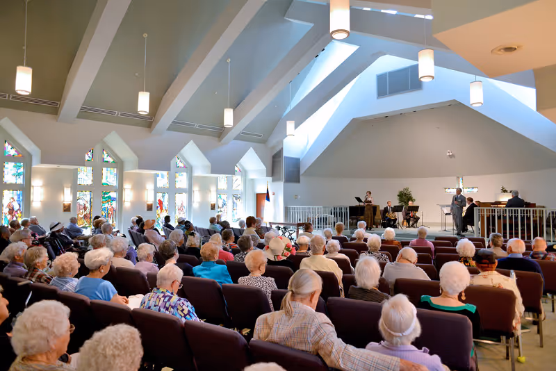 A large group of elderly people seated in rows of chairs inside a bright chapel or auditorium with high vaulted ceilings and stained glass windows. At the front, a few people are standing and sitting near a podium and musical instruments, possibly leading a service or event.