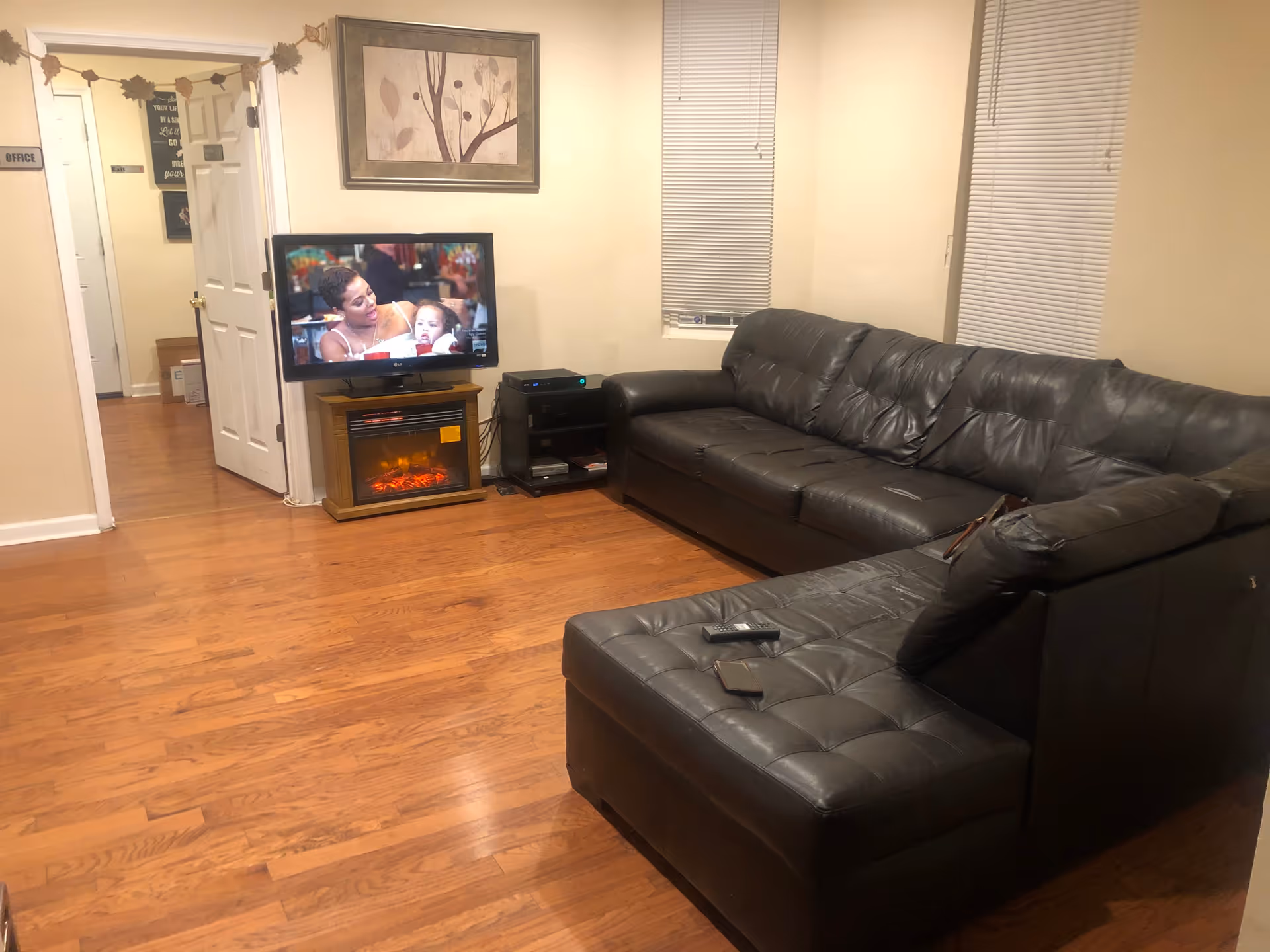 A living room with a large black leather sectional sofa, a flat-screen TV on a stand above an electric fireplace, hardwood floors, two windows with blinds, and a framed artwork on the wall. An open door leads to a hallway with a sign labeled 'Office'.