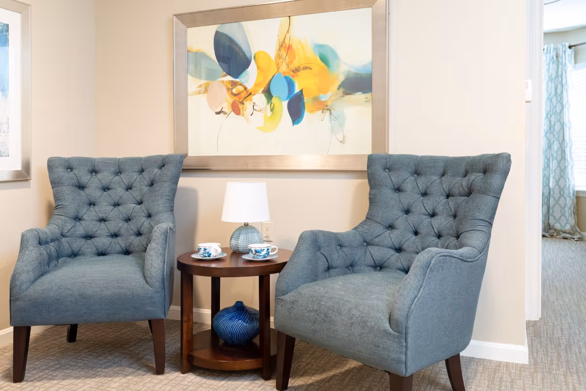 Two blue tufted armchairs with wooden legs are positioned on either side of a small round wooden table. On the table are two teacups with saucers and a small lamp with a white shade. Behind the chairs is a wall with a large framed abstract painting featuring blue, yellow, and orange colors. The room has beige walls and carpeted flooring, with a window partially visible on the right side with patterned curtains.