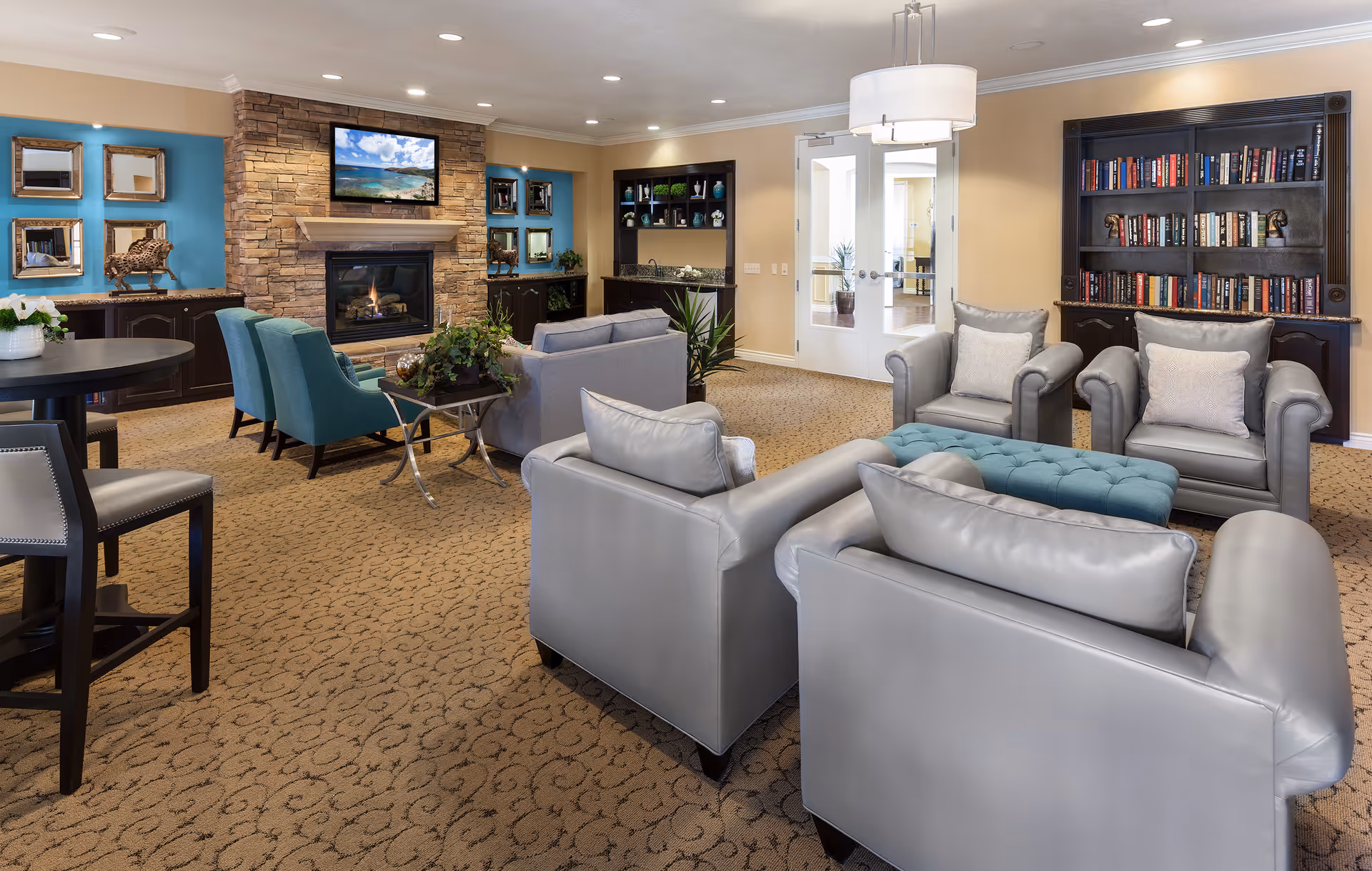 A cozy senior living facility common area with beige walls and carpet. The room features a stone fireplace with a TV mounted above it, two teal armchairs, two gray leather armchairs, a gray sofa, a blue tufted ottoman, and a dark wood bookshelf filled with books. There are decorative mirrors and sculptures on the walls and shelves, a small table with plants, and a modern ceiling light fixture. Double glass doors lead to another room.
