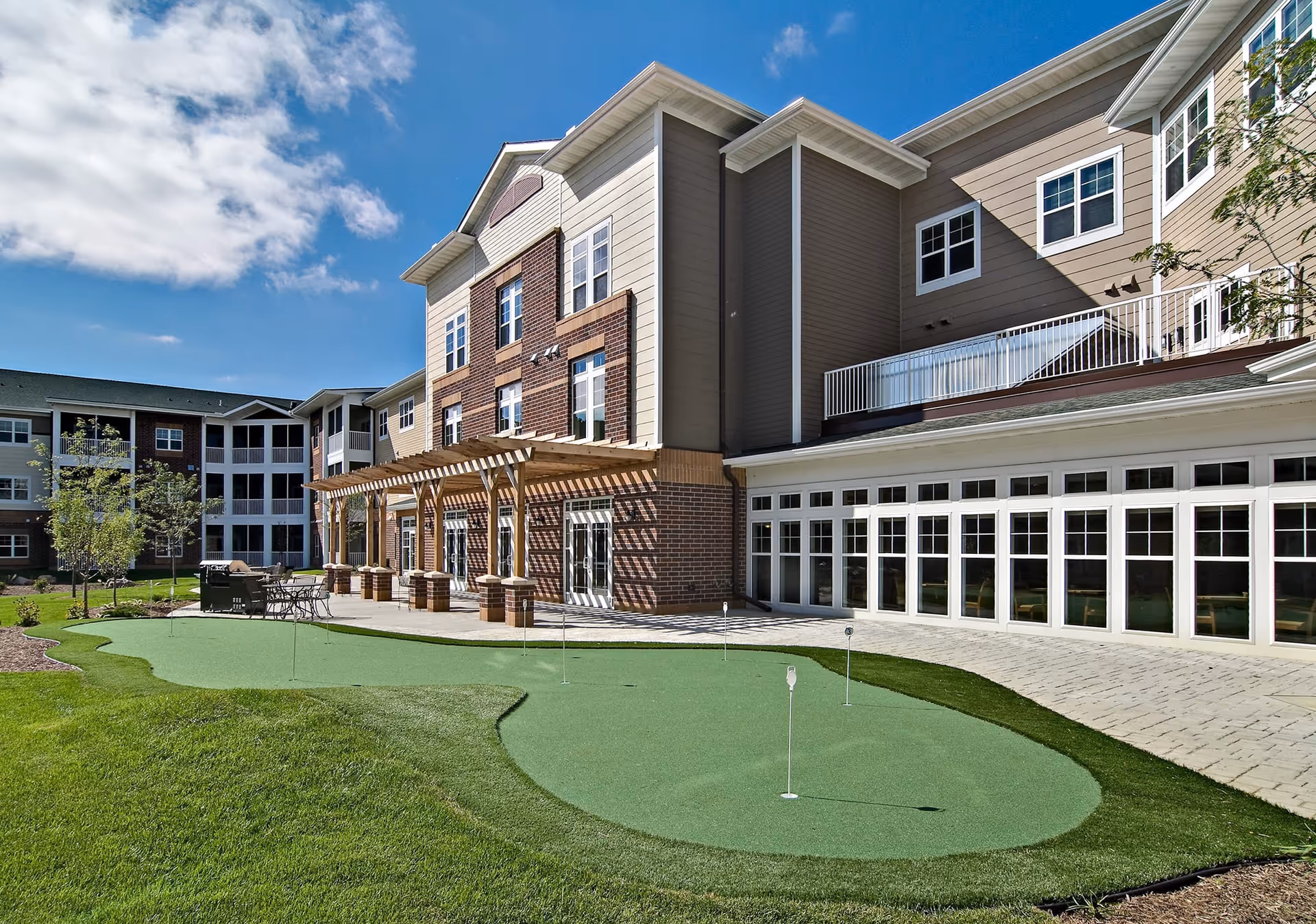 Exterior of a multi-story senior living building with a patio, pergola, and small putting green under a blue sky.