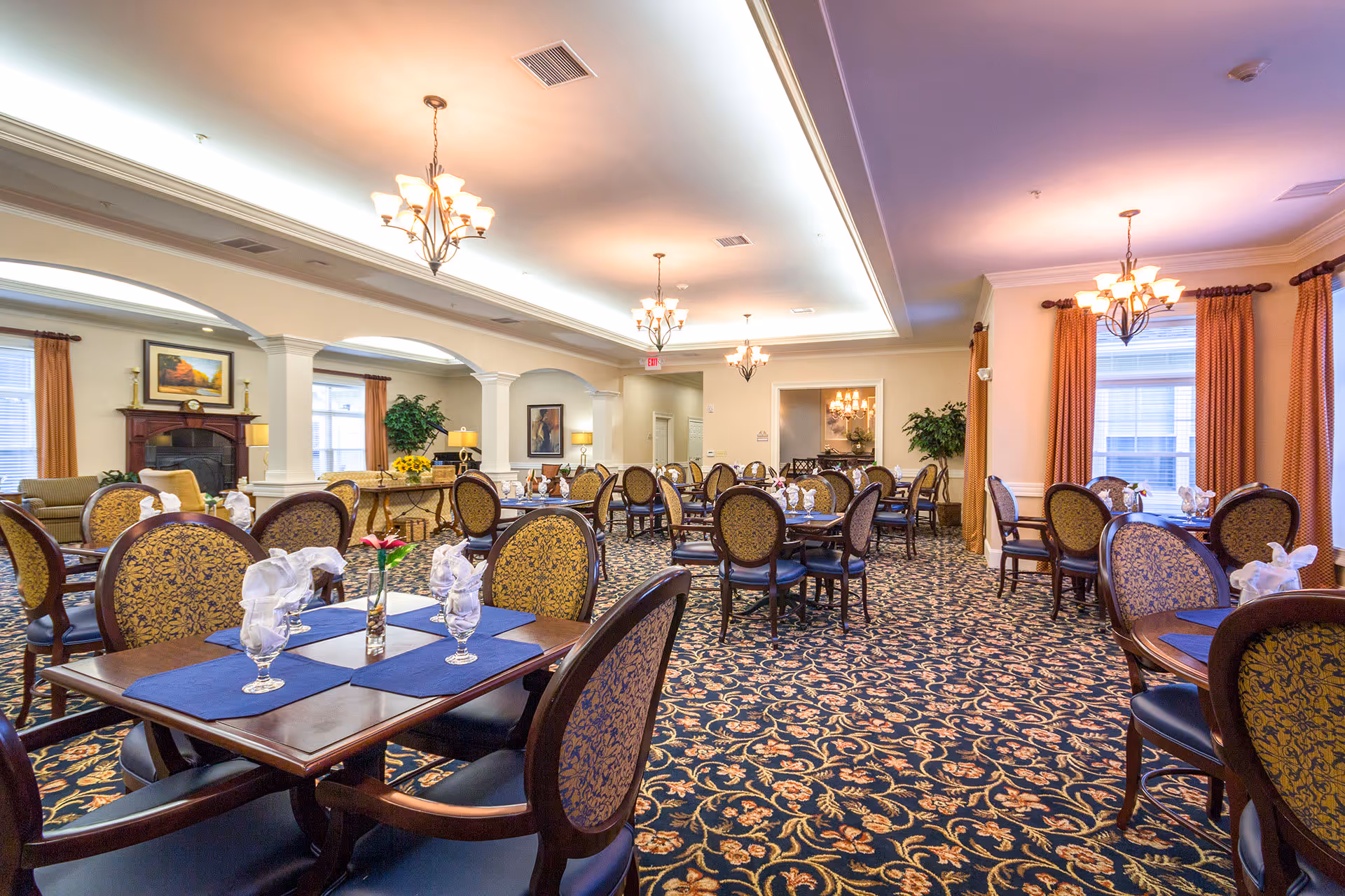 A spacious and elegant dining room with multiple round and rectangular tables set with blue placemats, glassware, and napkins. The room features patterned carpet, chandeliers, large windows with orange curtains, and decorative plants. There is a fireplace with a painting above it on one side of the room.