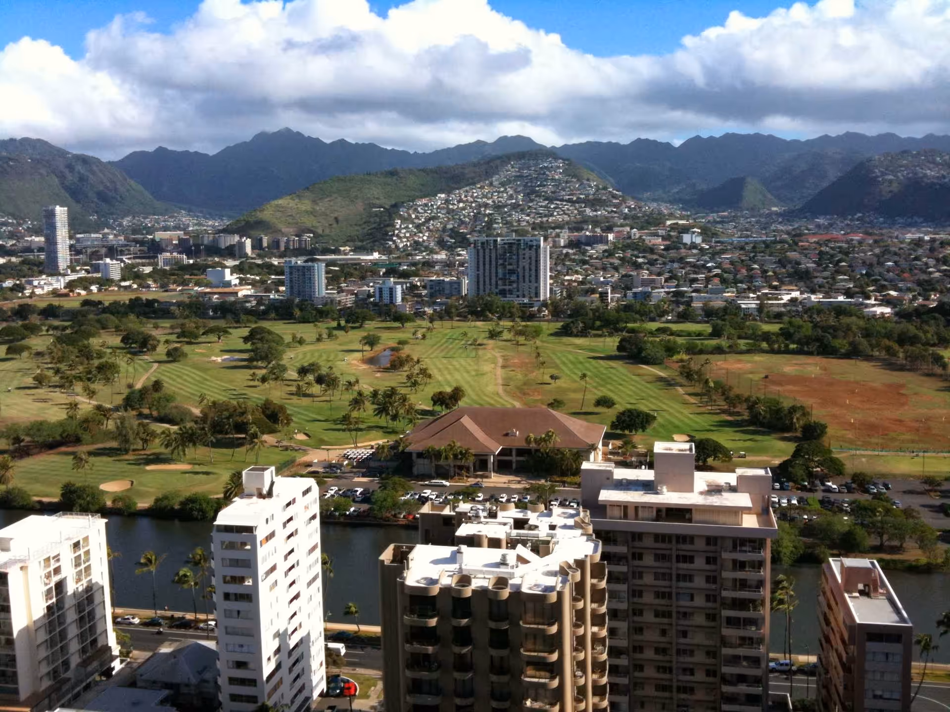 Aerial view of a cityscape with multiple high-rise buildings in the foreground, a golf course with green fairways and scattered trees in the midground, and a mountain range under a partly cloudy sky in the background.