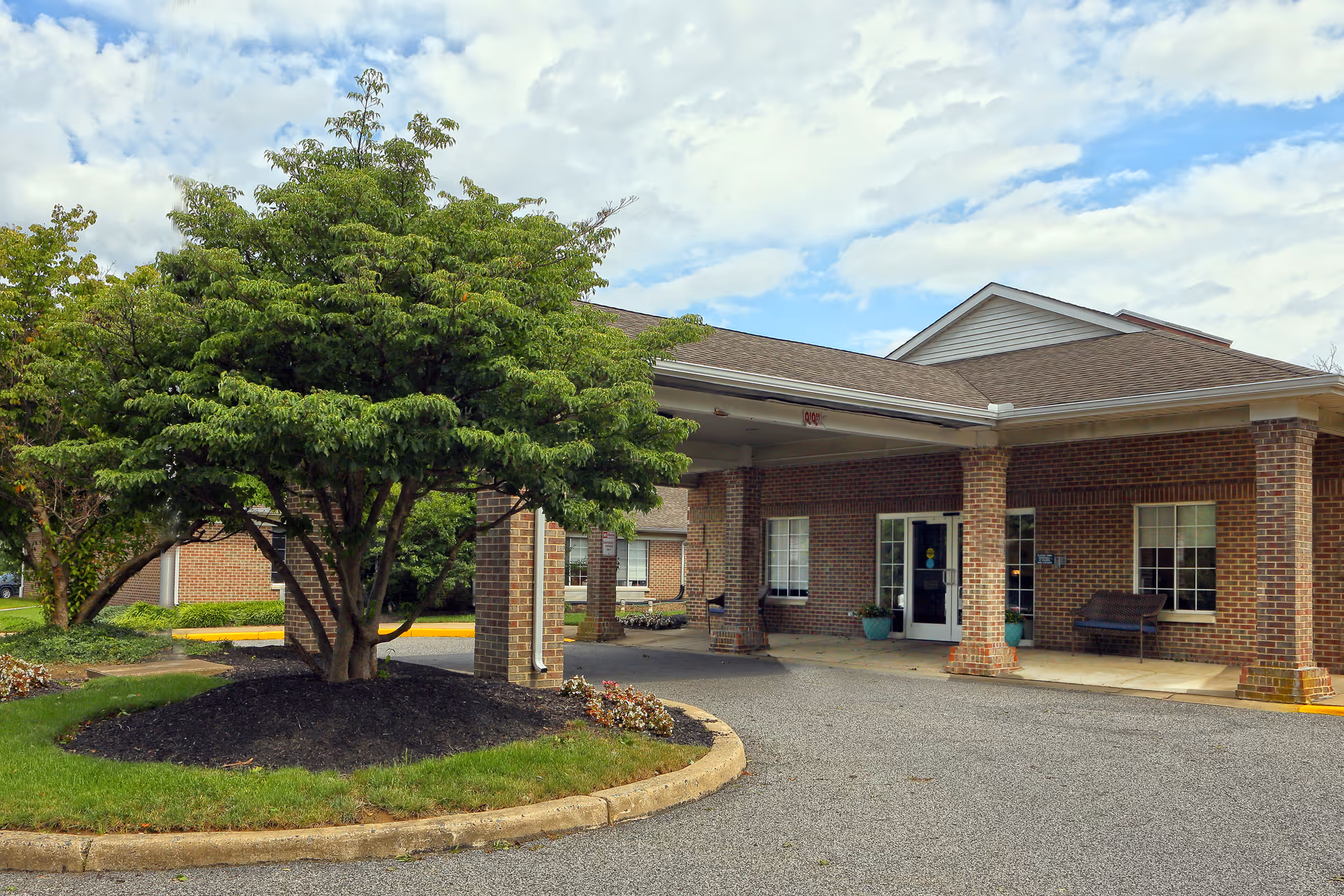 Exterior view of a brick building with a covered entrance supported by brick columns. There is a tree with green foliage in a mulched circular bed near the driveway. The sky is partly cloudy.