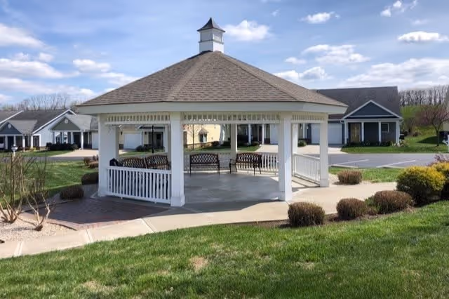 A white wooden gazebo with a shingled roof situated on a paved area surrounded by green grass and bushes. In the background, there are single-story buildings with dark roofs and white walls under a partly cloudy blue sky.