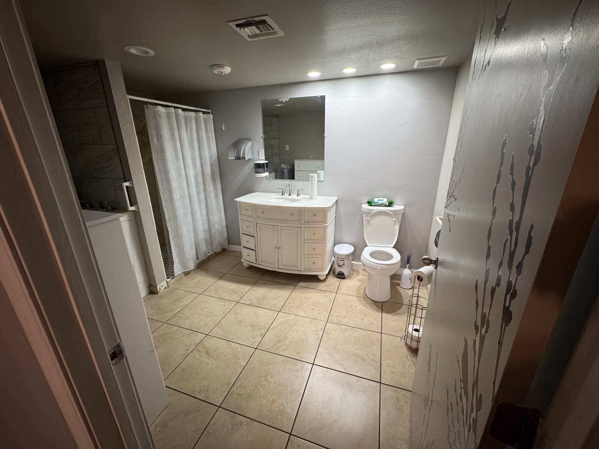 A clean bathroom with beige tiled floor, a white vanity with a sink and mirror above it, a toilet with the lid open, a shower area with a white curtain, and various bathroom accessories including a trash can, toilet brush, and paper towel holder.