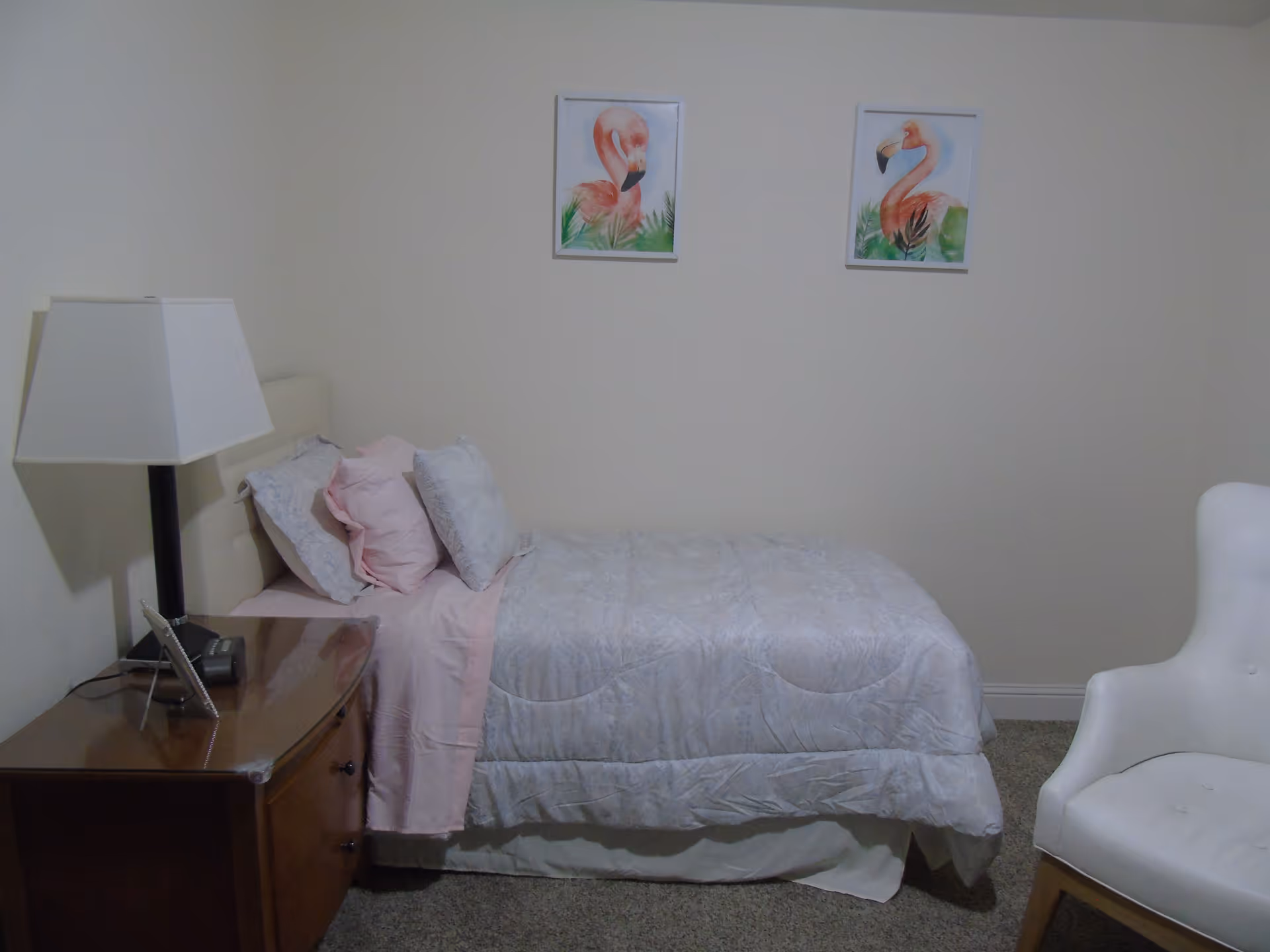 A small bedroom with a single bed, nightstand and lamp, a white armchair, and two flamingo prints on the wall.