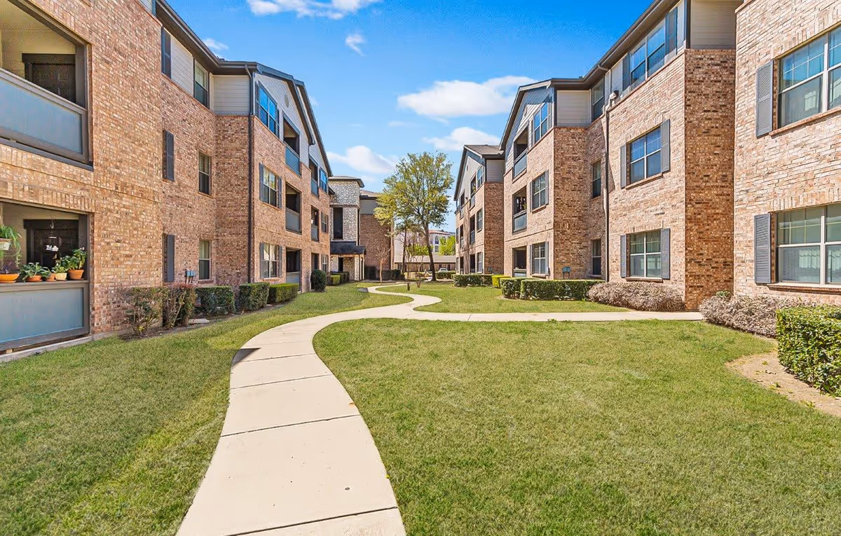 Outdoor view of The Cesera 55+ Apartments showing two three-story brick buildings facing each other with a curved concrete walkway and green lawn in between under a blue sky with some clouds.