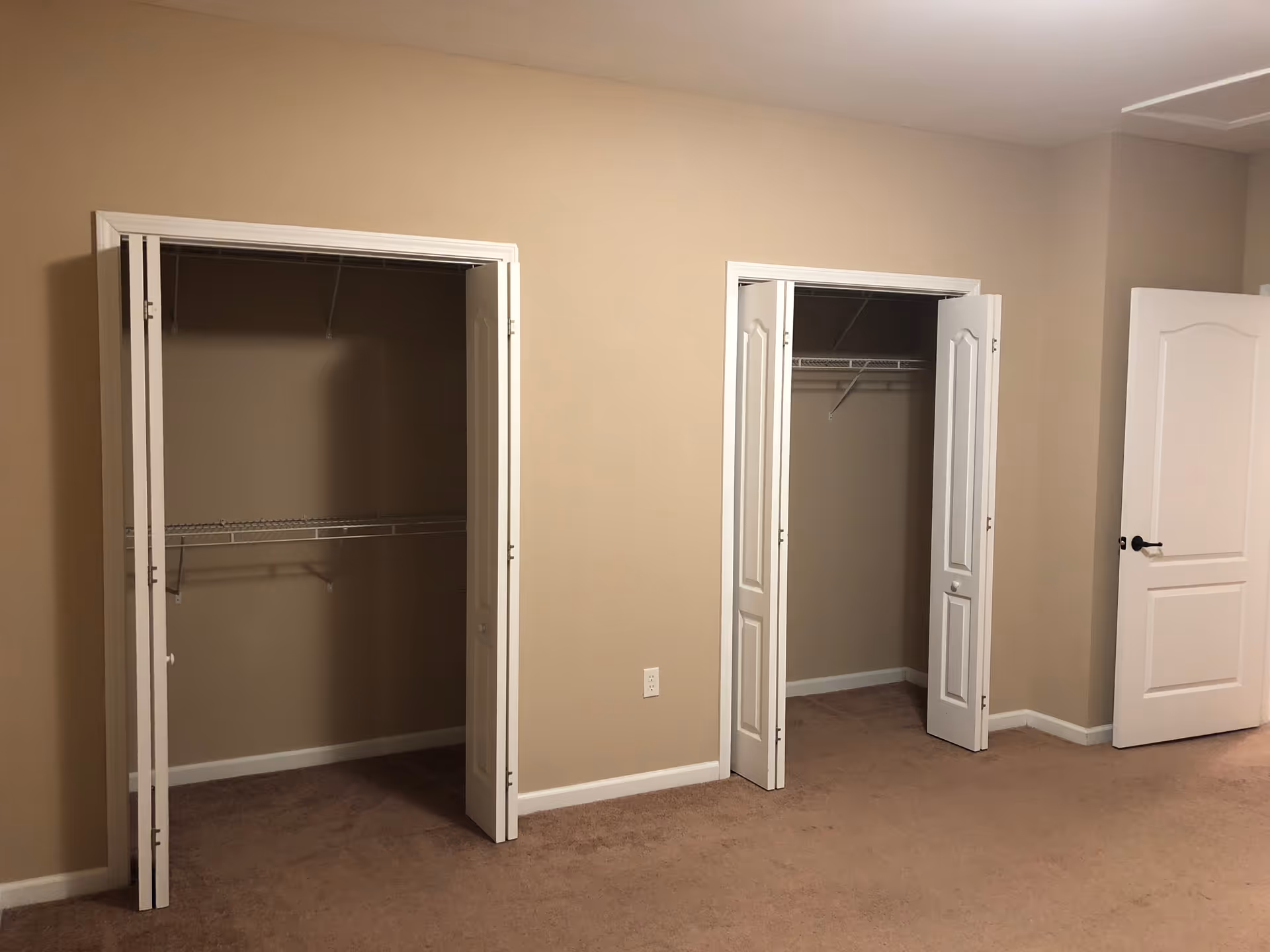 Interior view of a room with beige walls and carpeted floor featuring two open closets with white folding doors and a partially open white door to the right.