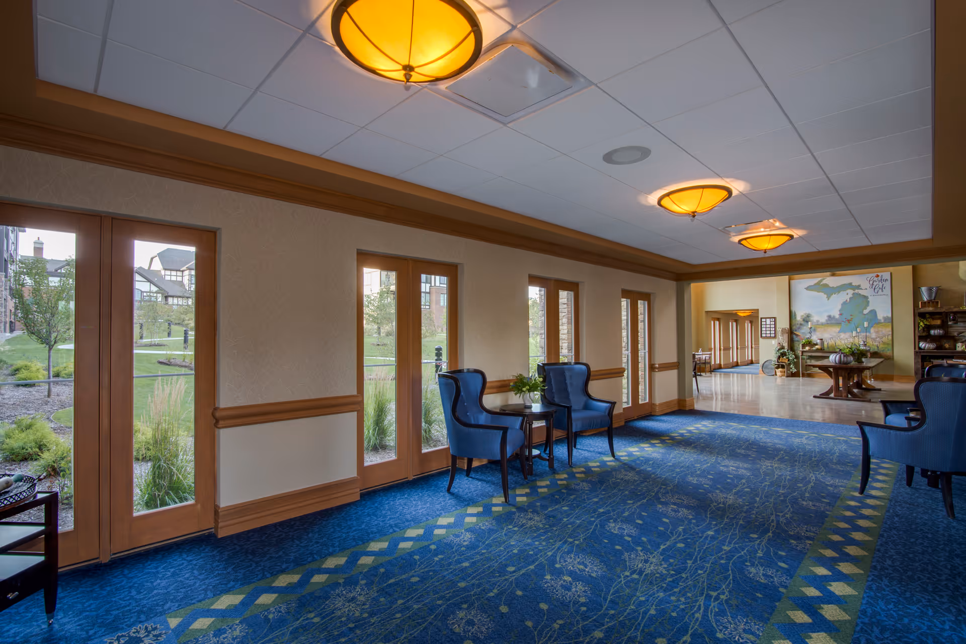 A spacious interior common area with blue carpet featuring a green and yellow pattern, several blue upholstered chairs paired with small round tables, and large windows and glass doors letting in natural light. The ceiling has recessed lighting fixtures, and there is a decorative table with plants and artwork visible in the background.