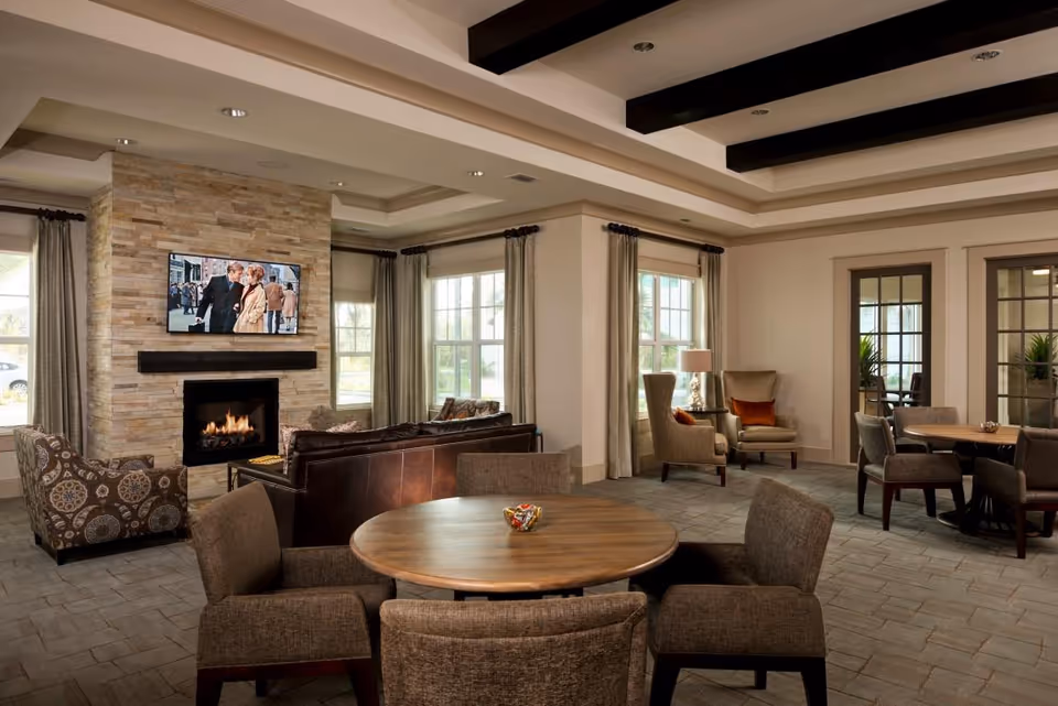 Spacious communal lounge with round tables and upholstered chairs, a stone fireplace with a mounted TV, and large windows letting in natural light.