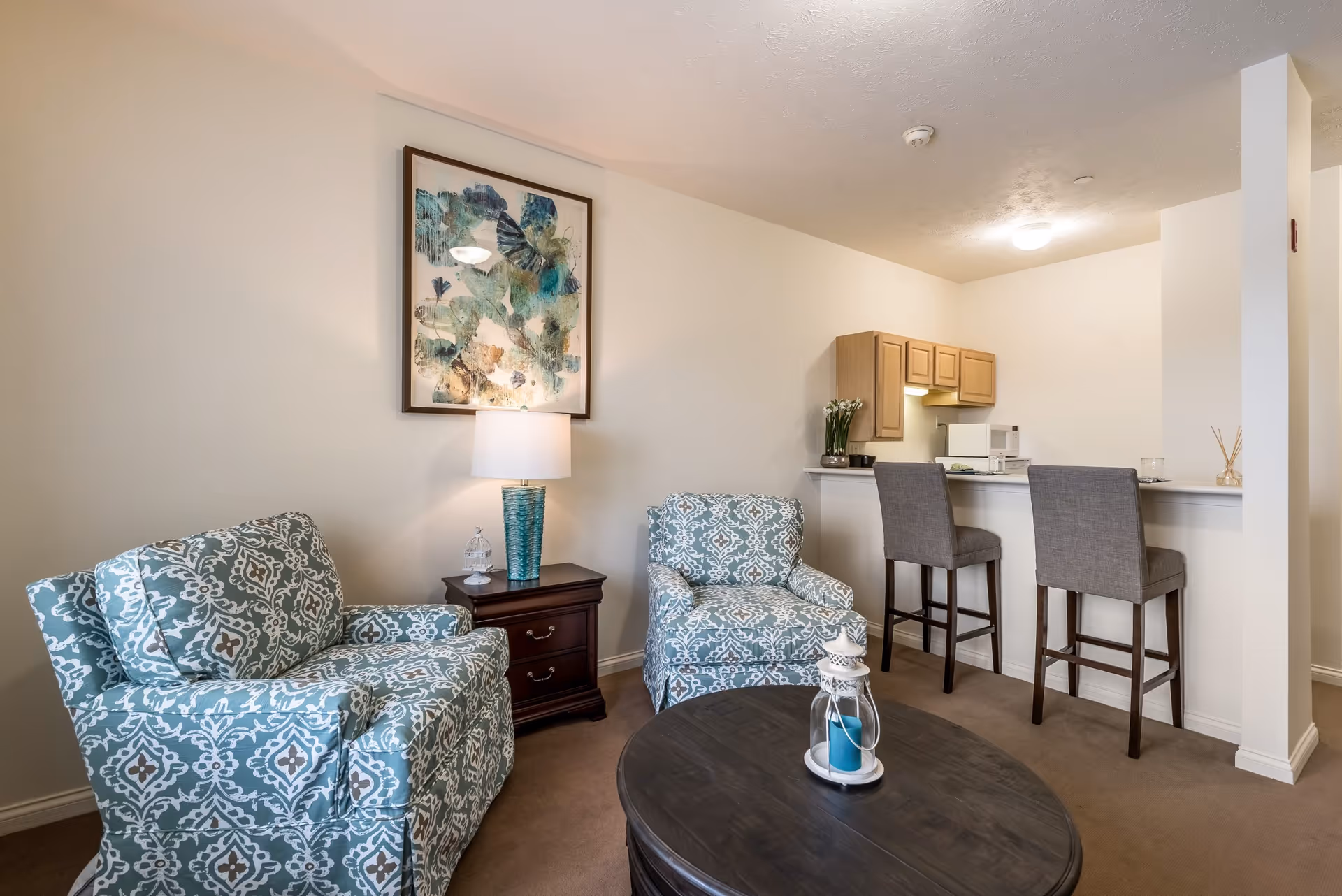 A cozy living area with two patterned armchairs in blue and white, a dark wooden coffee table with a decorative lantern, a small dark wooden side table with a lamp and a decorative item, and a kitchen counter with two gray bar stools. The kitchen area has light wood cabinets, a microwave, and a small plant on the counter. A framed abstract artwork hangs on the wall above the side table.