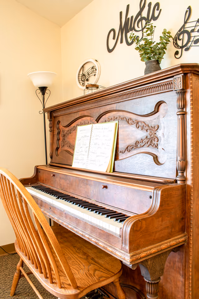 An ornate wooden upright piano with intricate carvings, an open sheet music book on the music stand, a wooden chair in front of the piano, a floor lamp to the left, and decorative wall art featuring musical notes and the word 'Music' above the piano.