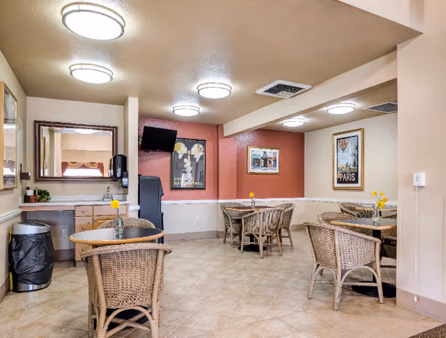 A cozy dining area in an assisted living facility with several round tables and wicker chairs. Each table has a small vase with yellow flowers. The walls are decorated with framed artwork, including a Paris poster. The room has tiled floors, beige walls, and ceiling lights. A trash can and a counter with a mirror and soap dispenser are visible on the left side.